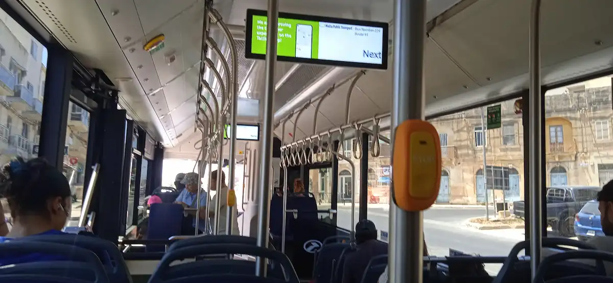 Interior de un autobús del transporte público en Malta, con pasajeros y pantallas de información de rutas.