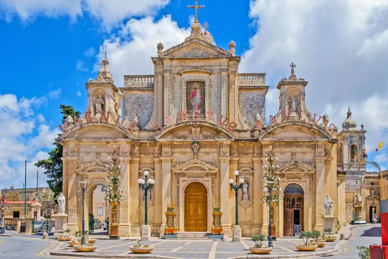Fachada barroca de la iglesia colegiata de San Pablo en Rabat, Malta, con estatuas de santos, columnas y decoración festiva bajo un cielo azul